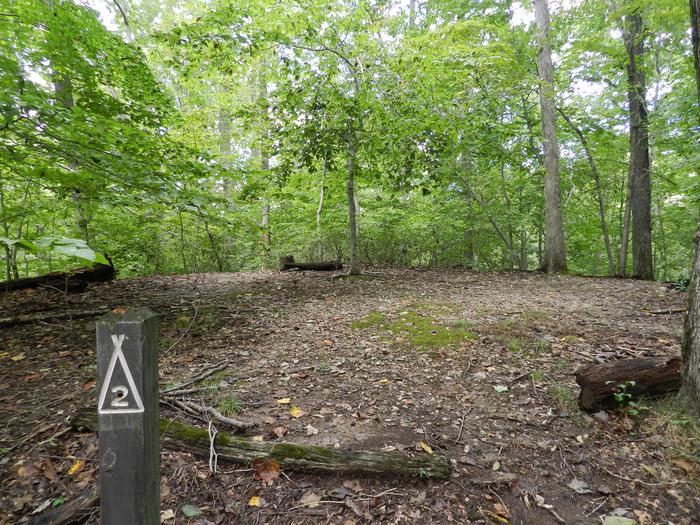 Area of cleared flat ground in the forest next to a wooden post labeled 2Site 2 is located up a short steep hill on a yellow blazed path to a knob.  This breezy location overlooks the water and is cooler and less buggy in summer.