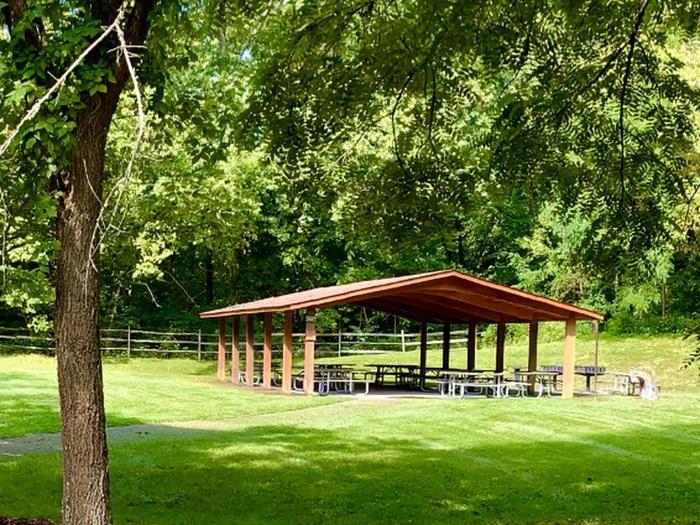 Brown open-aired shelter surrounded by mature trees and green grass. Inside the shelter are numerous picnic tables and grills available for picnic use.Sycamore Picnic Shelter