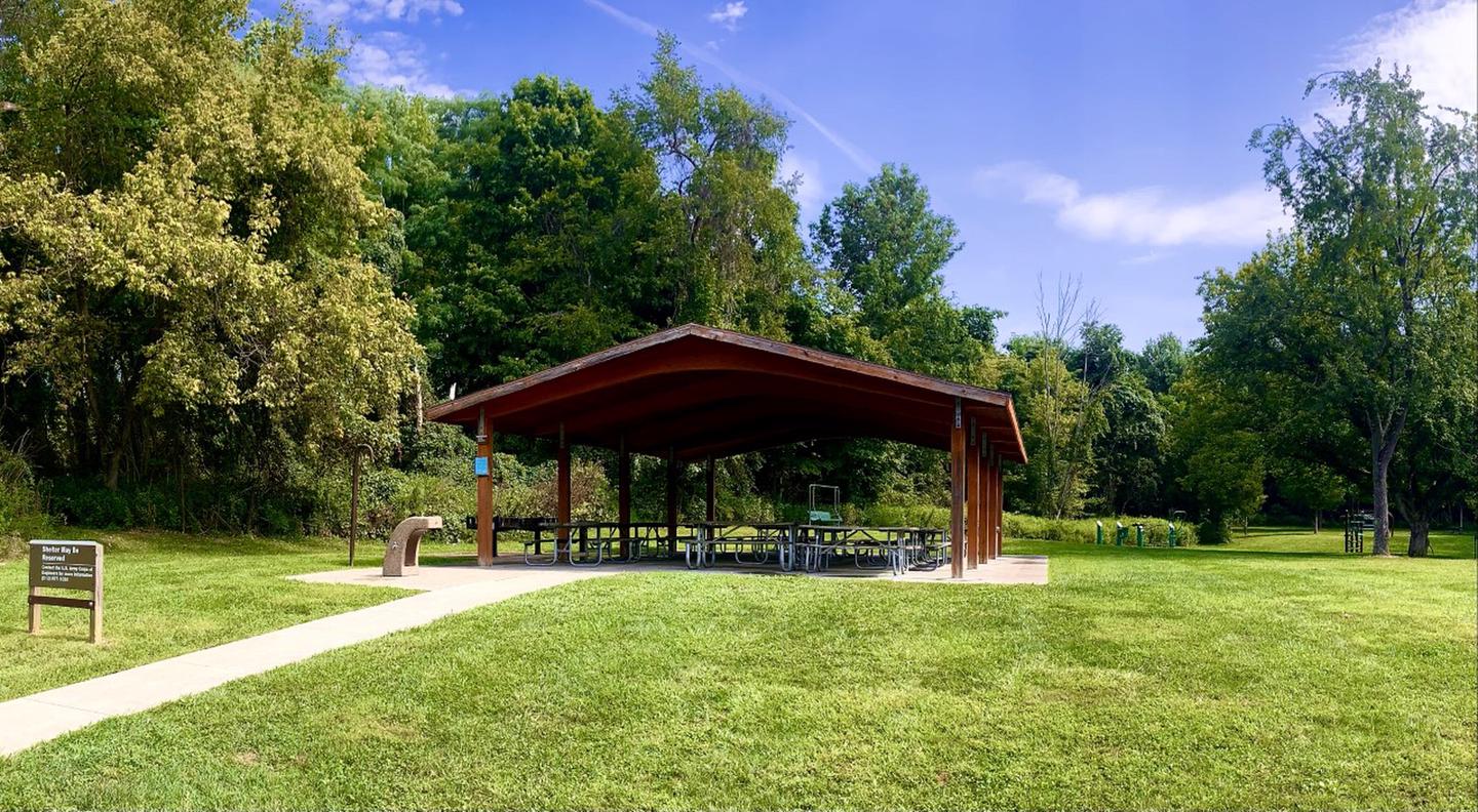 Open-sided brown shelter surrounded by mature trees and a green lawn.Holly Picnic Shelter view from parking lot