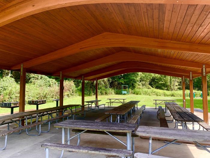 Inside view of a light brown wooden open-aired shelter. Various picnic tables, grills, and a swing are also present in the vicinity. Inside of Holly Picnic Shelter