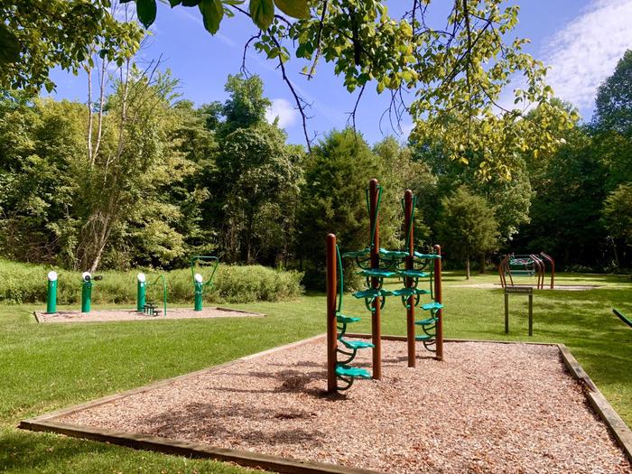 Fitness playground surrounded by green grass and mature trees. 4 climbing posts and outside workout equipment are present.Fitness playground located behind Holly Picnic Shelter