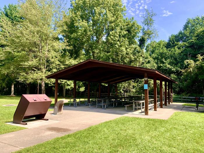 Brown wooden shelter surrounded by mature trees and green grassRedbud Picnic Shelter