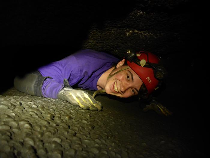 A person in caving gear crawls through a tight cave passage.The tightest squeeze is 8.5" tall by 24" wide.