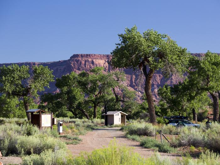 A registration kiosk and and vault toilet under Cottonwood trees with red rock cliffs in the background.Creek Pasture campground