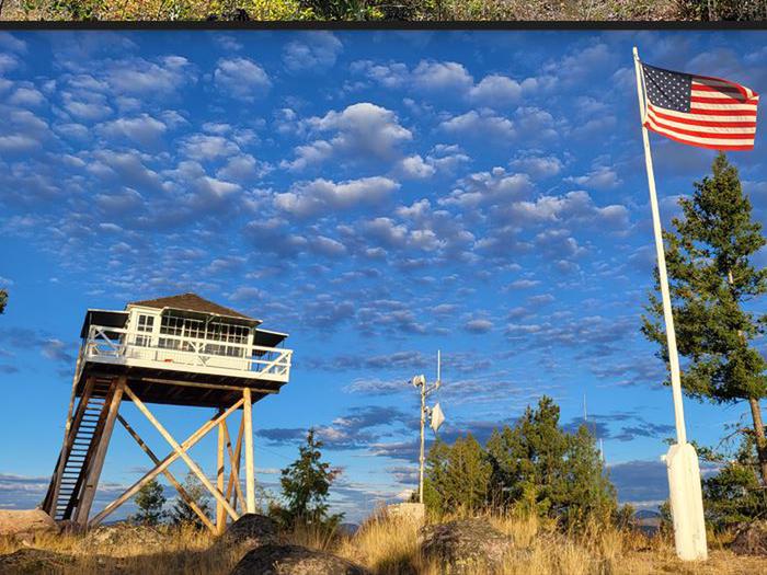 In background Double Arrow Fire Lookout tower with USA flag on flag pole in foreground with beautiful blue ski and light fluffy clouds passing byLookout Tower facing West