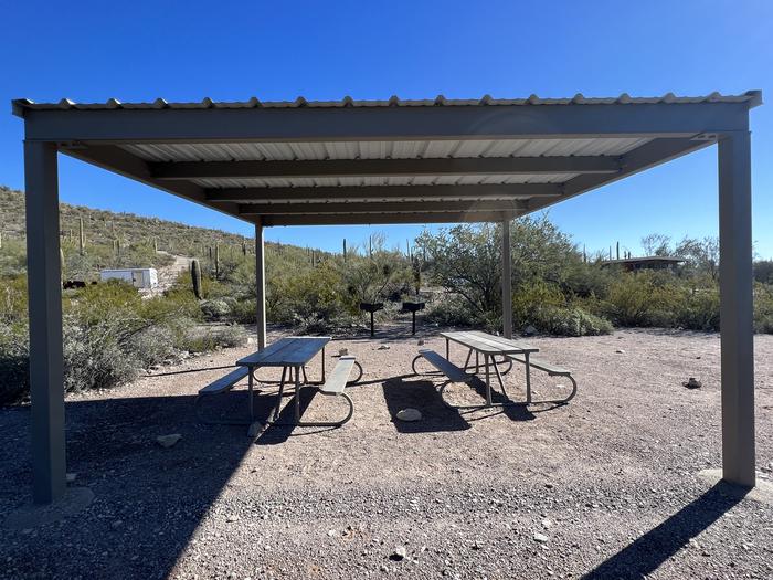 Picnic tables sit near grills and desert vegetation with a shade shelter.Each site has a picnic table and grill.