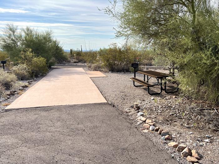 Pull-thru campsite with picnic table and grill, cactus and desert vegetation surround site.  The entrance into Site 090