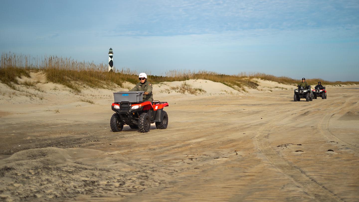Three ATV are driving on the beach, with the dunes behind them. The Cape Lookout Lighthouse is shown in the top left. Three ATV riders drive down the beach on South Core Banks