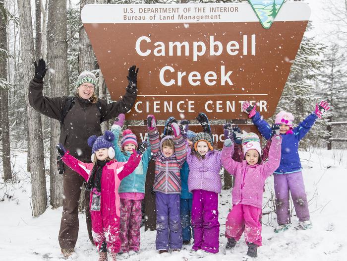 Students and an instructor raise their hands above their heads in celebration in front of a sign that says Campbell Creek Science Center. Students explore the snow at Campbell Creek Science Center. 