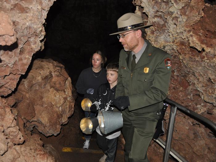 A park ranger and two children hold candle buckets while walking through a dark cave.Candlelight Tour participants must be 8 years and older and carry a candle bucket for light.