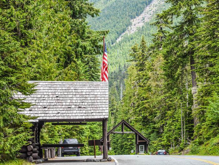 White River Entrance Station with wooden cabins on a highway with one building flying an American flagA tree-lined highway with two wooden buildings on the side of the road, one flying an American flag at the White River entrance. Tall conifer trees line the road. A mountain looms in the distance.
