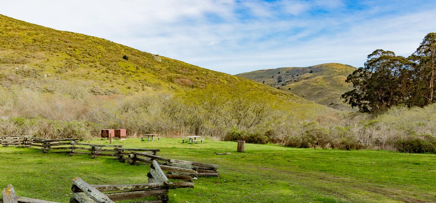 An image of a grassy meadow 2 picnic tables and bear lockers, and green hills in the distance.Site 1 at Haypress Campground in springtime. 