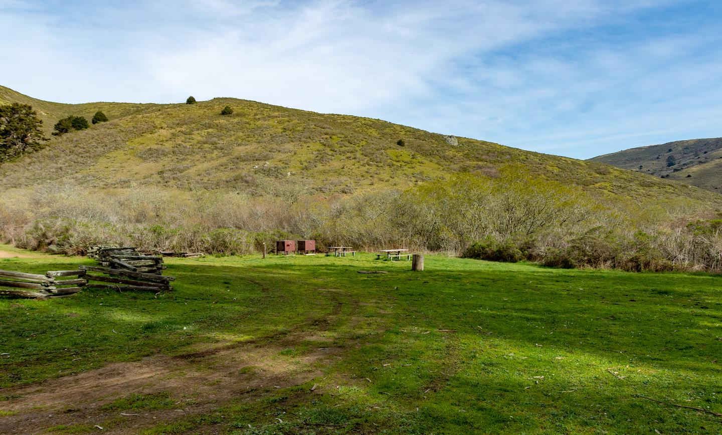 Big grassy field with bear lockers and picnic tables. Shrub covered hills in the distance.Wide view of Haypress Group Site.