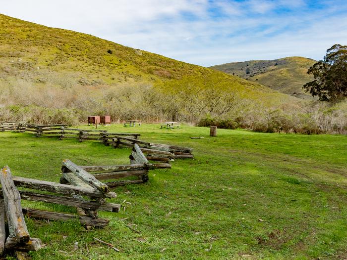  A low log fence runs along a grassy field. There are two picnic tables and two food lockers. In the distance are rolling, shrub-covered hills.Group site at Haypress Campground.