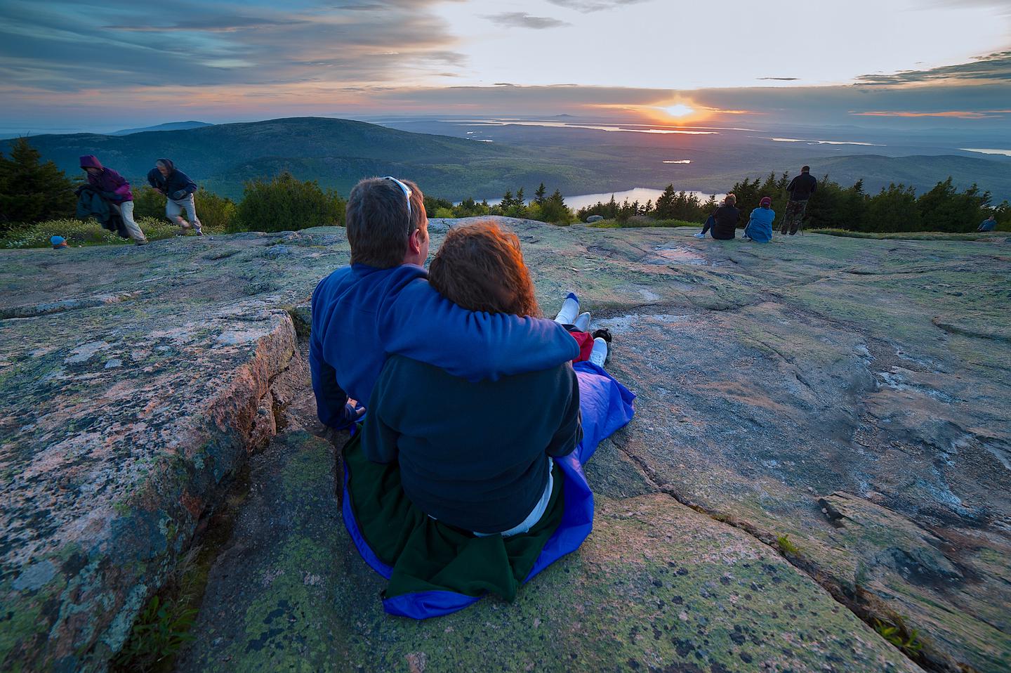 Male and female couple sitting on rock slab with arms around each other overlooking a sunset across a distant lake.Sunset view from Cadillac Mountain.