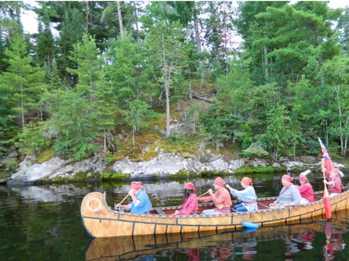 Seven park visitors paddle a 26-foot long north canoe on a lake with rocky shorelines in the backgroundPark visitors enjoy learning about the life of a Voyageur aboard a 26-foot north canoe.