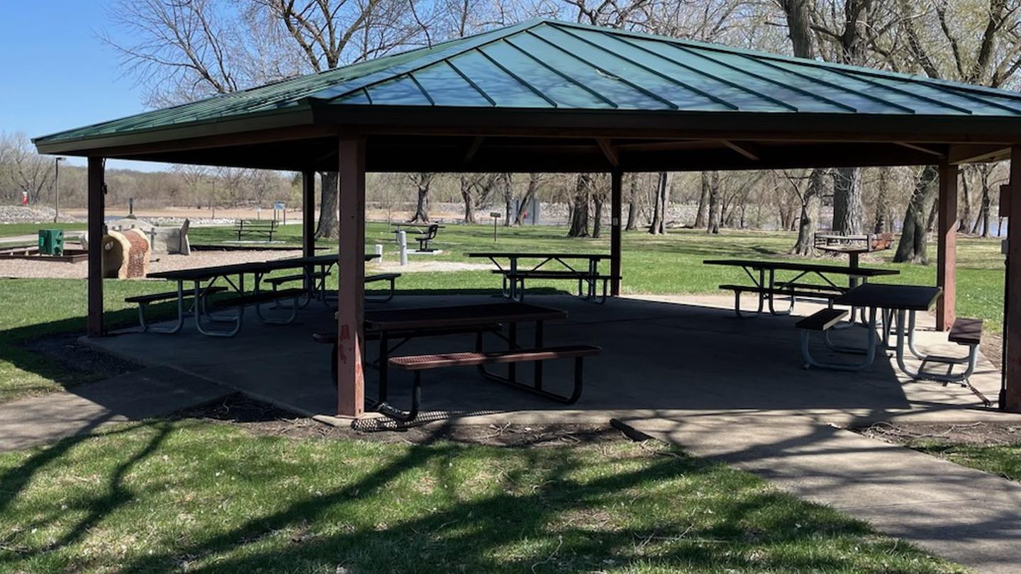 Large park shelter with picnic tables and grill. View of playground and park benches in the background. South Tailwater Shelter