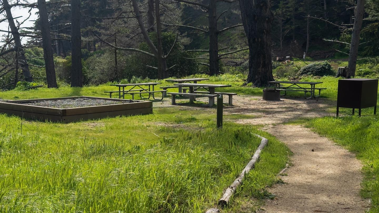 A sign post with "2" on it designating the entrance to Site 2. There are picnic tables, a fire pit, bear locker, and in the foreground is an elevated tent pad for ADA Access.Site 2 of Kirby Cove.