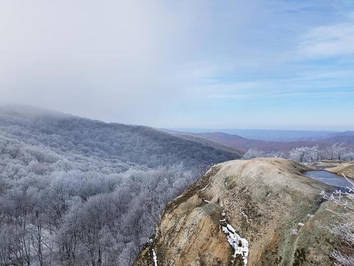 A grayish-white, rounded rock cliff in front of a scenic view of forested mountains white with snow.White Rocks offers a challenging hike with magnificent scenic rewards.