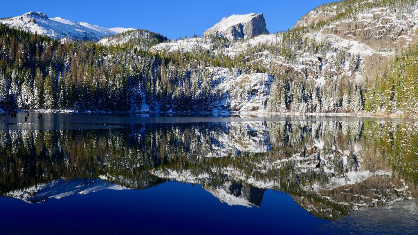 View of Bear Lake in spring. There is snow and ice on the mountains and trees surrounding the lake. There is a reflection of Hallett Peak in Bear Lake.Bear Lake in spring