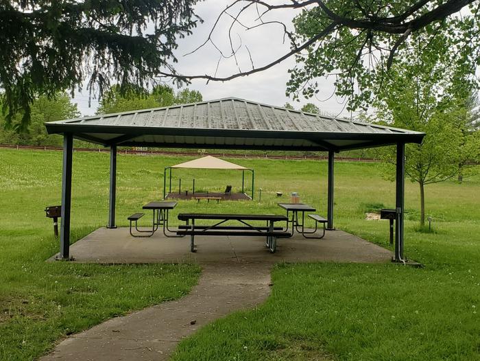 Front Side of shelter house in grassy area with trees. There are 3 picnic tables, two standing grills, and electrical outlet on the right hand side.TBD