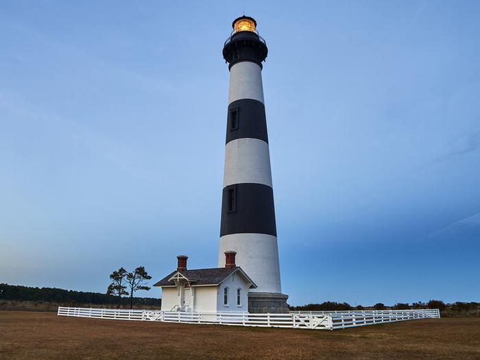 Bodie Island lighthouse at Sunset showing surrounding landscape with adjacent oil house and fence surrounding the lighthouse base.Sunset at Bodie Island Lighthouse