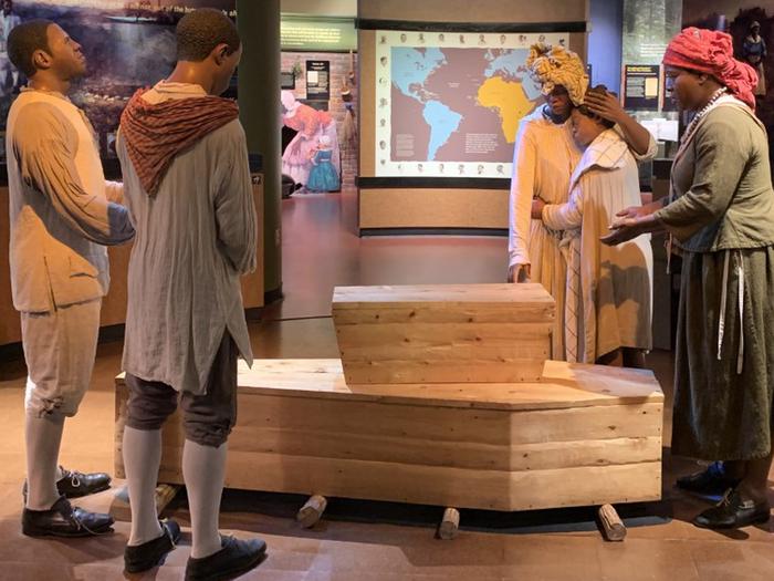 Five people of African descent wear colonial clothes and stand around an adult-sized coffin with a child-sized coffin on top of the adult-sized coffin.The burial scene at the African Burial Ground National Monument Visitor Center. 