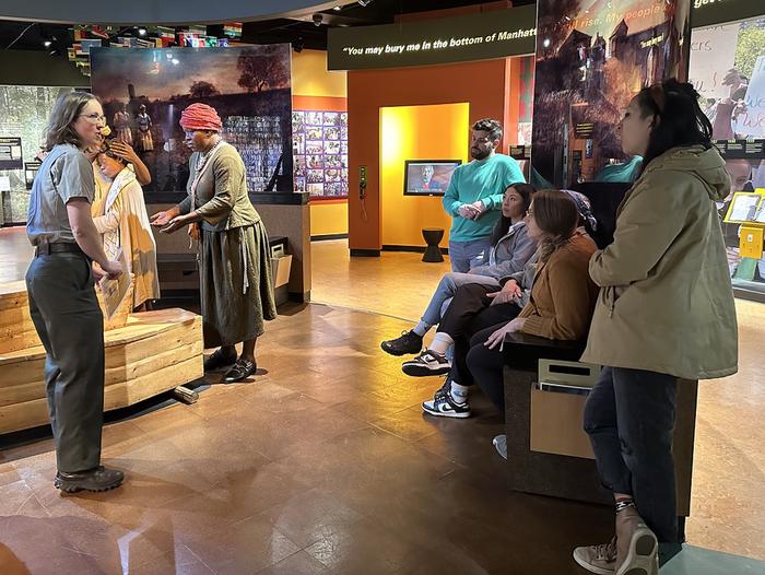 A ranger gives a tour to three visitors who are seated and two visitors who are standing. Museum exhibits are in the background. Ranger Bethany gives a tour to visitors at the African Burial Ground National Monument