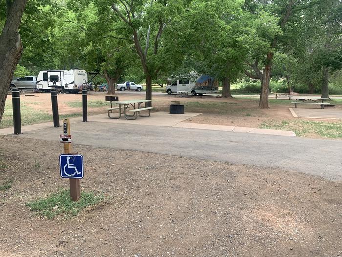 A paved driveway. Facing the end of the driveway, three bollards are in the ground at the end of the site spaced far enough apart for a bicycle to get through. To the right side there is a picnic table, grill ,and raised firepit all on pavement. A sidewalk leads from the picnic table. To the left side of the driveway there is a post with the blue accessible sign on it. There are many trees in the background.Site 26, Loop B in summer. Accessible site.
Paved Dimensions: 16' x 29'