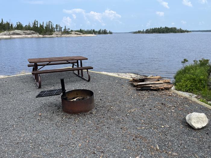 View looking out from campsite with the fire ring, picnic table, and stack of wood with water in the background.View looking out from campsite.
