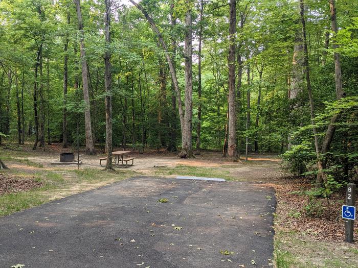 Wide paved handicapped parking space, picnic table, and fire ring in a shaded forest campsiteCampsite B2