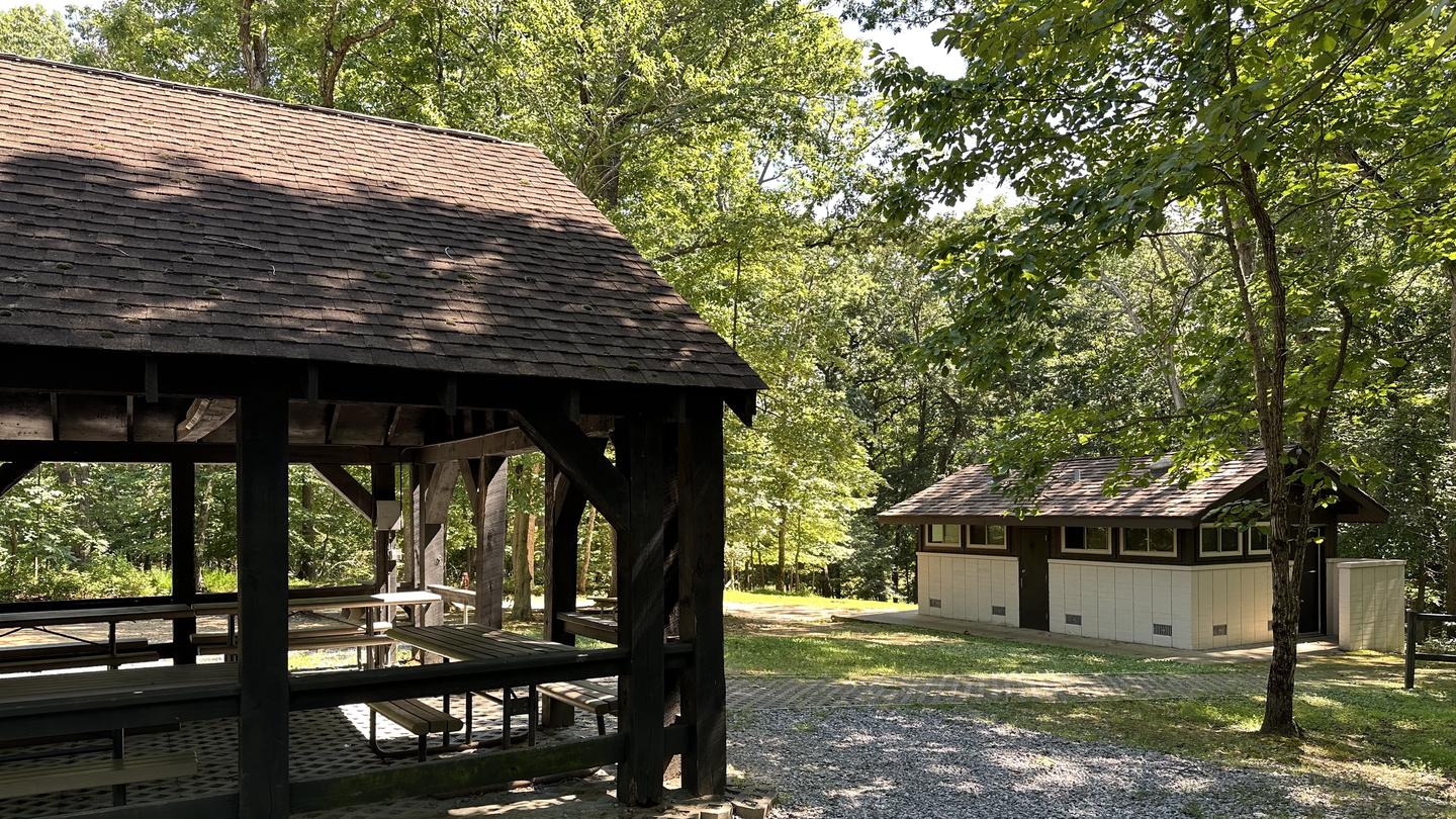 Open sided wooden picnic pavilion sits in a grassy area with a sidewalk leading to a cement restroom building in the forestTelegraph Picnic Pavilion and restrooms on a sunny day