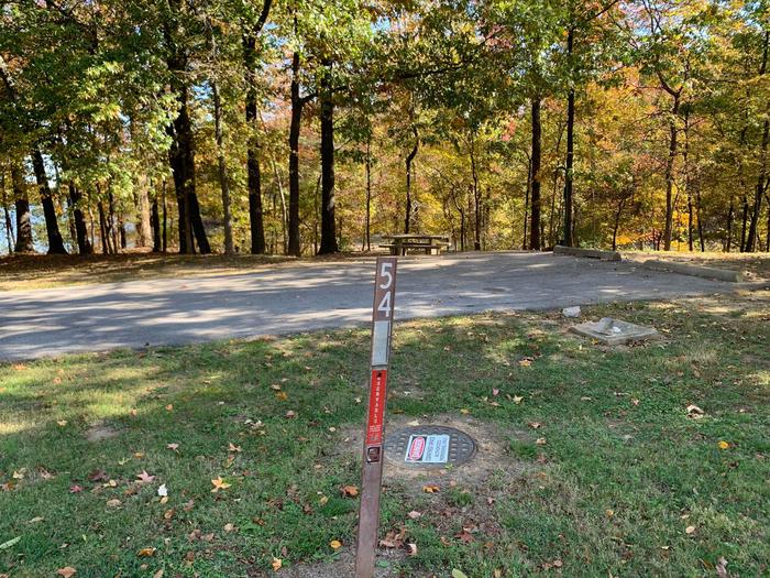 This site is nestled in the trees providing shade. The picnic table and fire pit are to the left of the concrete pad. The picnic table sits on a paved pad. This is a full hookup site and all hookups are located on the right. 