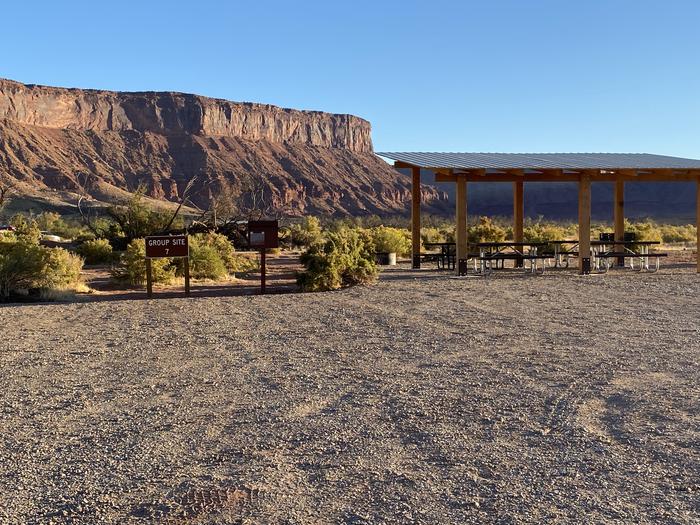 The group site parking area, shade structure, and picnic tables with scenic red rock cliffs in the background.Group campsite 7 has a shade structure with picnic tables, a standing grill, and firepit. It is in an open area with views of the surrounding red rock cliffs and the banks of the Colorado River.