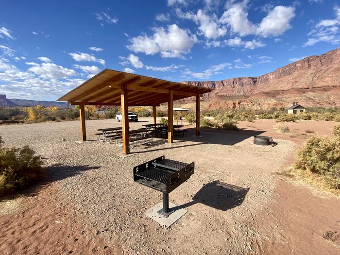 The shade shelter with four picnic tables underneath, adjacent firepit, and barbeque with the river and high cliffs in the background.Group campsite 7 has a shade structure with picnic tables, a standing grill, and firepit. It is in an open area with views of the surrounding red rock cliffs and the banks of the Colorado River.