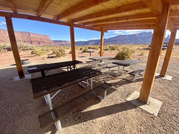 Closeup view of four average sized picnic tables underneath a wooden shade structure.Group campsite 7 has a shade structure with picnic tables, a standing grill, and firepit. It is in an open area with views of the surrounding red rock cliffs and the banks of the Colorado River.
