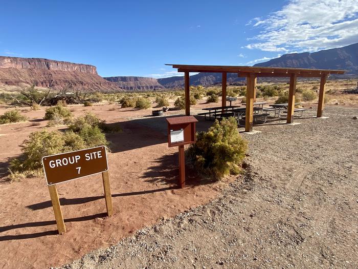The sign and bulletin board for campsite 7 in the foreground with the site behind them and a view toward the upper Colorado River canyon with high cliffs. Group campsite 7 has a shade structure with picnic tables, a standing grill, and firepit. It is in an open area with views of the surrounding red rock cliffs and the banks of the Colorado River.