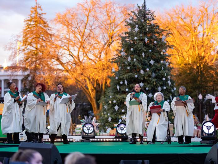 Six women in white robes stand on the stage and singThe Mt. Calvary Catholic Church Choir performs at the 2022 National Christmas Tree Lighting