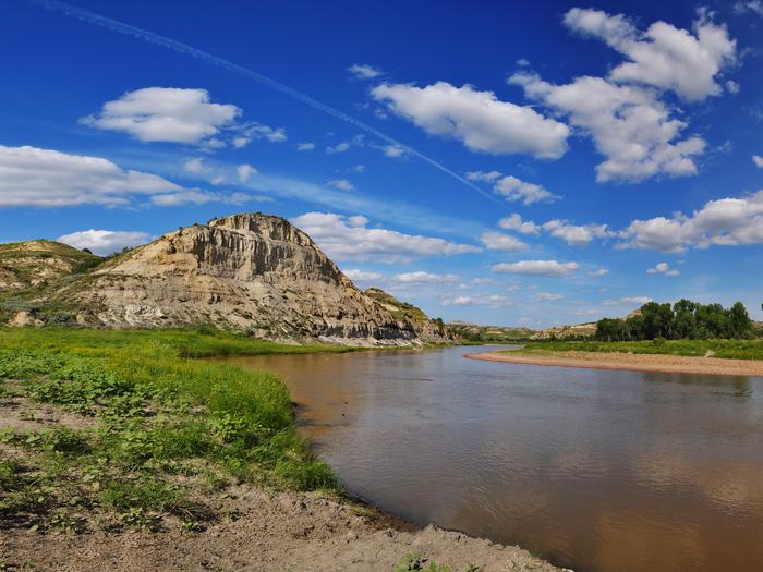 The Little Missouri River flows under a deep blue sky with a dramatic butte in the backgroundThe Little Missouri River under a deep blue sky with a dramatic butte in the background