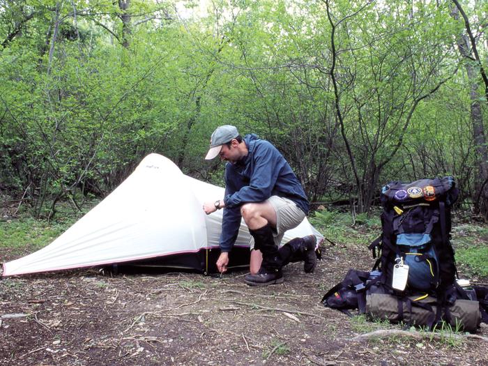 Person wearing hat, raincoat, and shorts setting up small white tent in the forest with a backpack next to themBackcountry camper setting up tent.