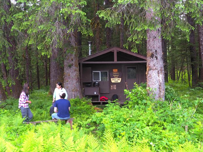Three people outside brown cabin with trees and fernsEagle Lake cabin
