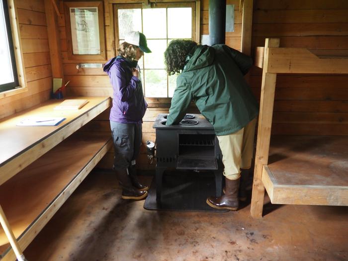 Two people lighting the stove in Gut Island Cabin 2 with wooden bunkbeds and cabinetsGut Island 2 interior