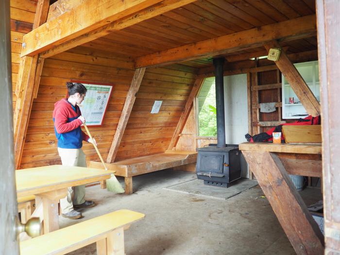 Person sweeping the interior of wood cabin with wood table and bunkbed with a black stoveLittle Dry Island Cabin interior