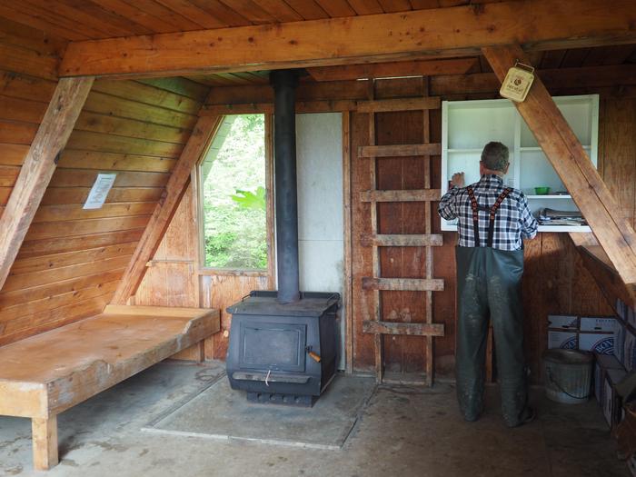 Man at shelves inside wood cabin with black stove and wood bunkInterior of Little Dry Island Cabin