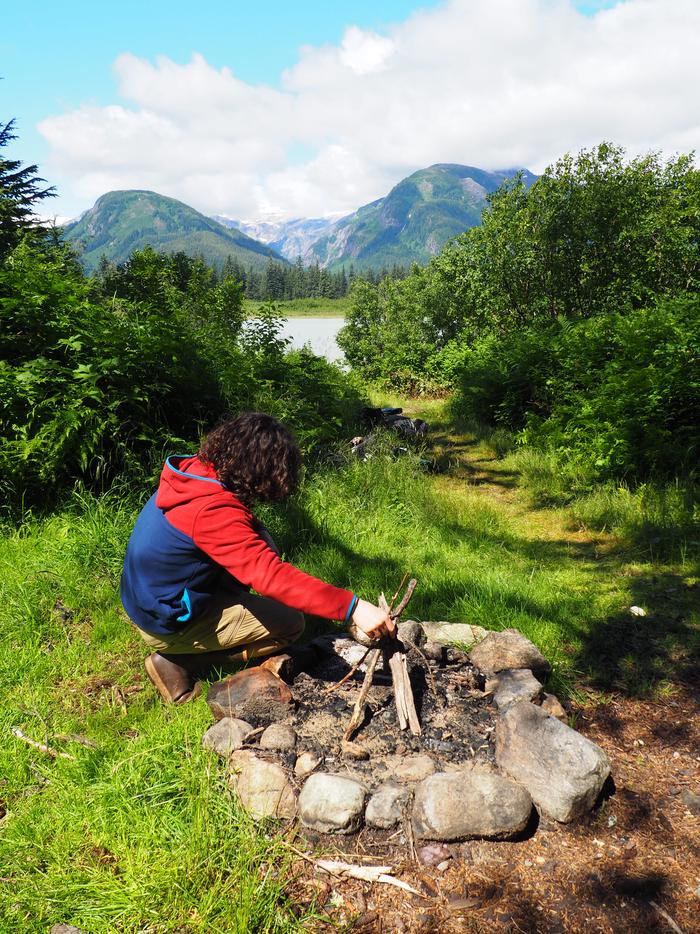 A person building a teepee of sticks in a rock firepit with mountains is backgroundBuilding a fire in front of Shakes Slough 1 Cabin 