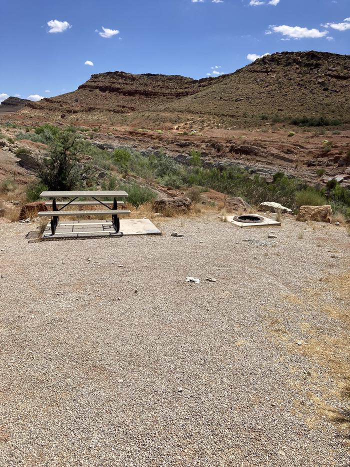 Gravel camping pad and picnic table at Quail Creek Reservoir Designated Dispersed Camping AreaRolling hills with striated, red rock formations surround a flat, gravel camping pad with associated picnic table and metal fire ring.   
