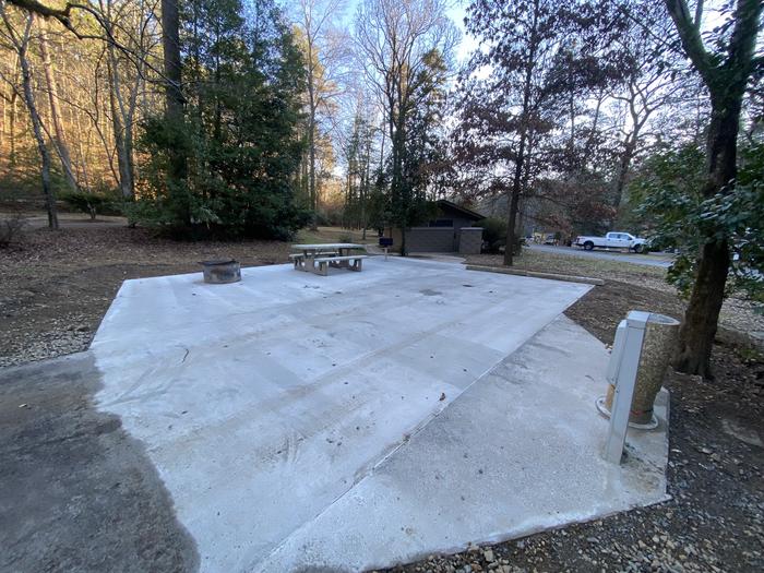 A white concrete pad with a fire ring, hook ups, and picnic table sit beneath a canopy of trees. A view of Site 4. The parking pad, picnic area, grill, and fire ring are all surrounded by concrete. A smooth paved path leads to the comfort station. 