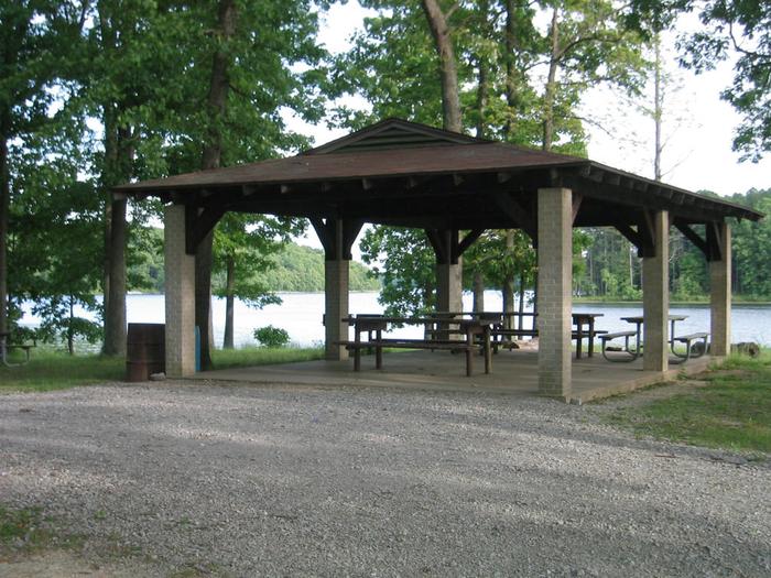 A pavilion with picnic tables, concrete floor and brick columns. The pavilion is next to a lake and shaded by trees. A gravel road connects to the pavilion.Devils Kitchen Group Pavilion
