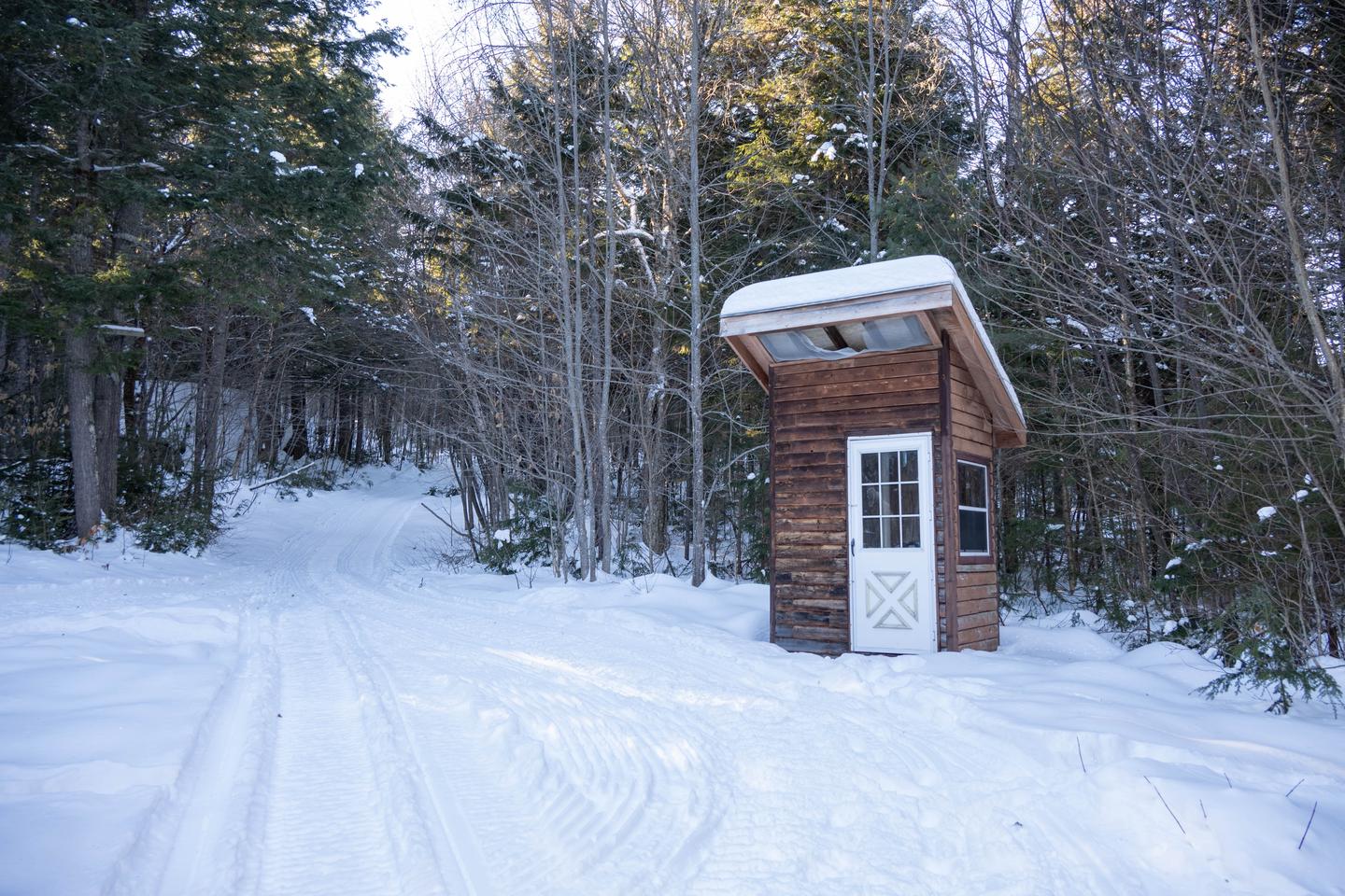 A small rectangular wooden shack structure with a white door and slanted roof is the structure of a pit toilet. The building is outside surrounded by snow in the woods.An outhouse is available just outside of Big Spring Brook Hut.