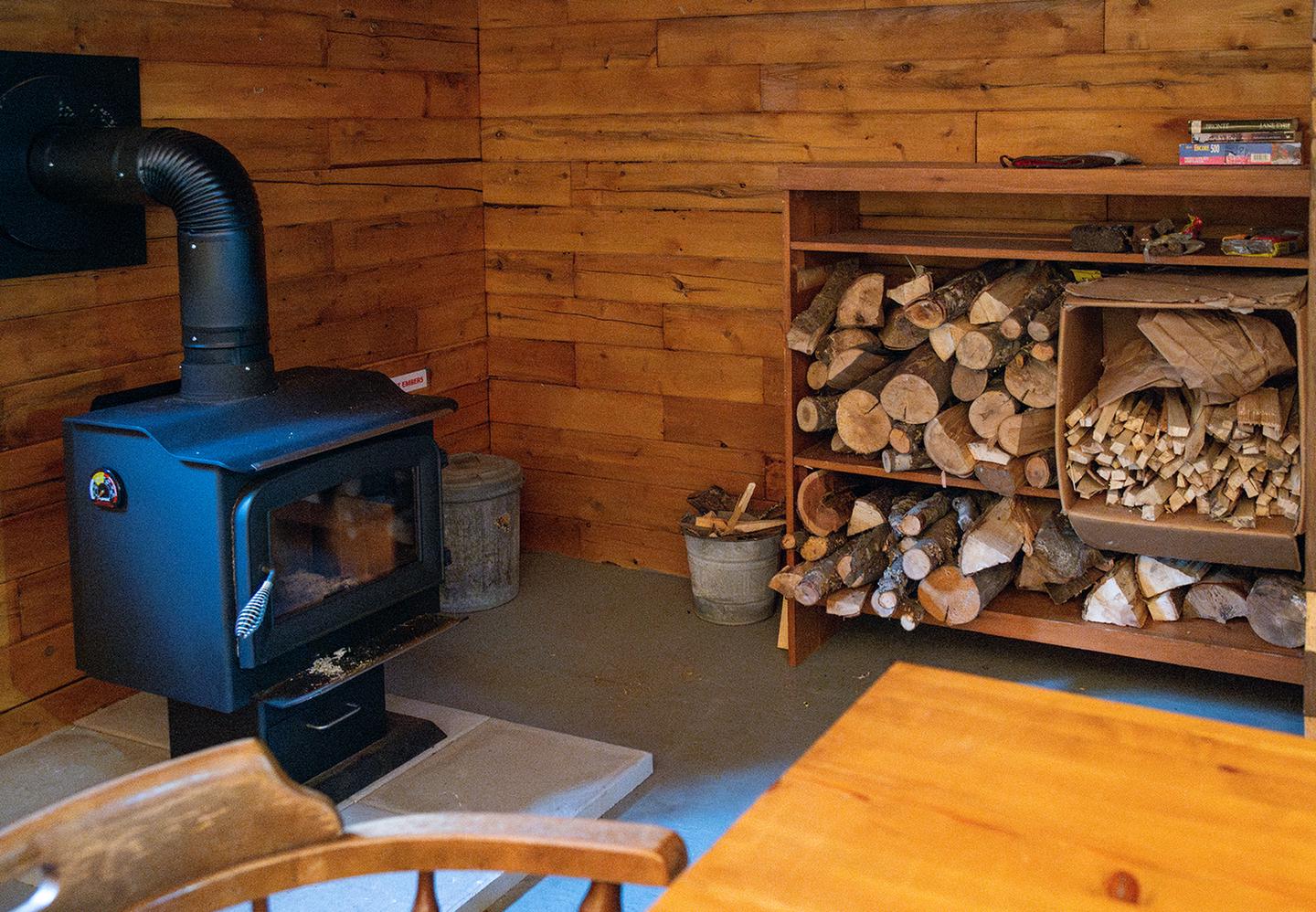 A black woodstove on the left and neatly arranged firewood to the right, adjacent to the stove sit inside of a wooden cabin.Stay warm by the woodstove in Big Spring Brook Hut.